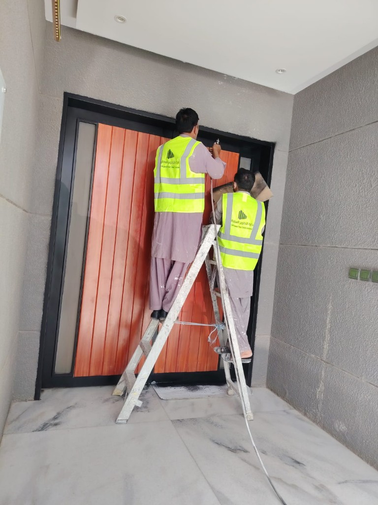 الصيانة الدورية للعقارات في الرياض: ماذا يفعل الفريق قبل أن يرنّ هاتف المستأجر؟ فنيان من دارة التطوير على سلم أثناء صيانة إطار باب خشبي لمدخل مبنى سكني حديث بالرياض