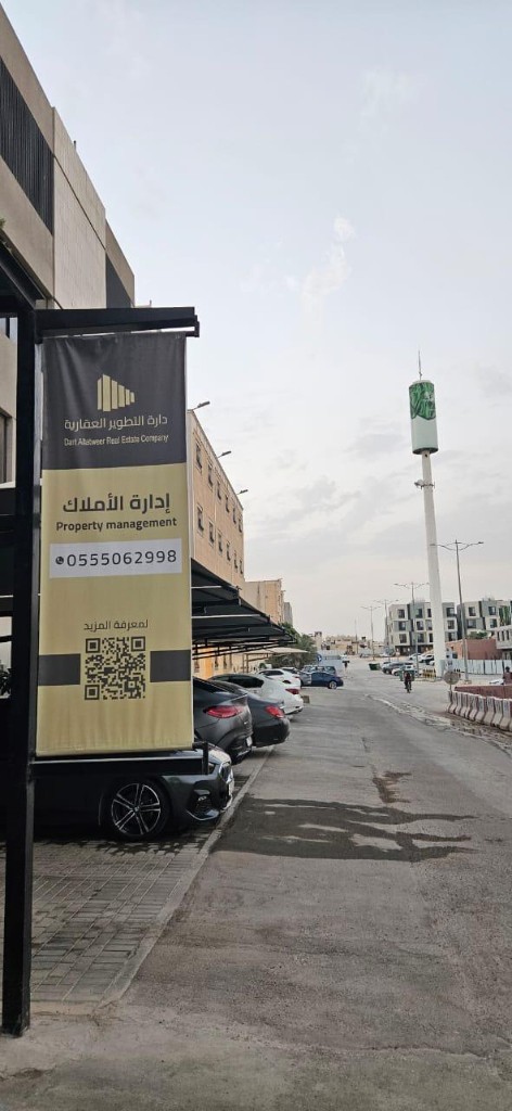 إدارة أملاك عقارية على ثلاث واجهات: اللافتة والمقر والممر بنر دعائي لدارة التطوير عن إدارة الأملاك مع رمز QR في موقف سيارات بالرياض